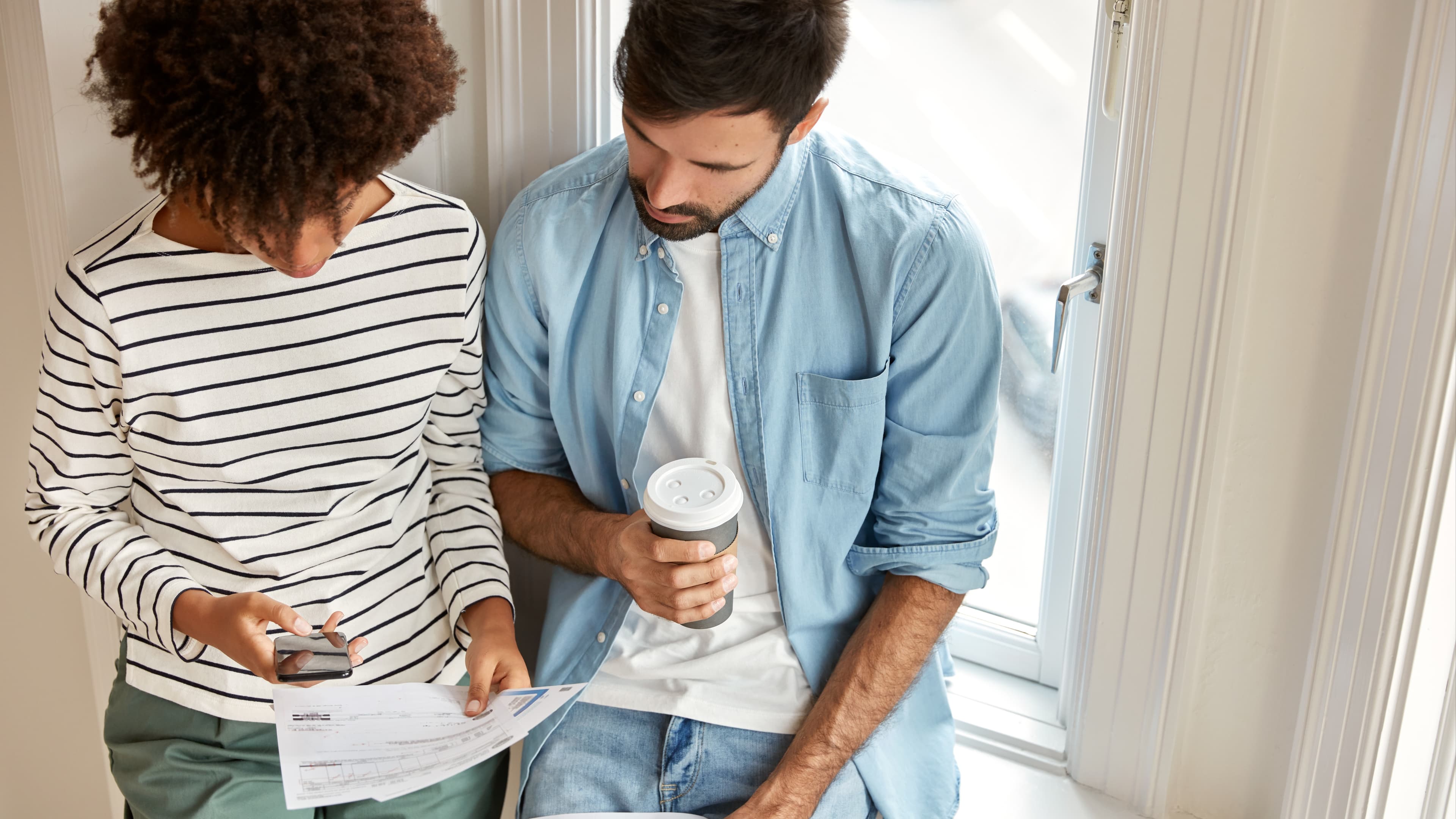 Top view of multiethnic managers discuss marketing report, work together, check information on mobile phone drink takeaway coffee stand against window wall. Unshaven man and his assistant