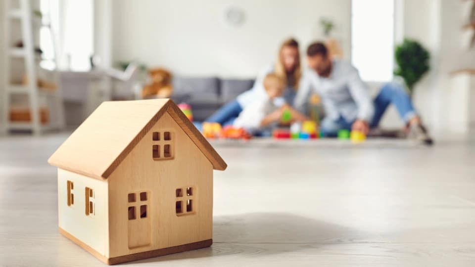 Close up of miniature detached house on floor at home with happy family playing with child in the background