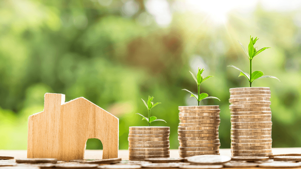 Stack of gold coins next to a small miniature wooden house